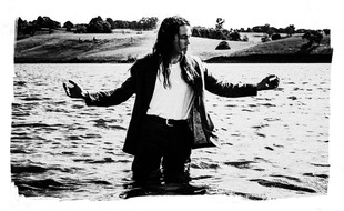 A worn, black & white photo of Didirri standing in a lake with the water up to his thighs. He is wearing black pants with a white tucked t-shirt, a belt, and a black long sleeved over-shirt. In the background are grassy hills and trees. He has his arms outstretched to the sides, slightly bent, and looks to his left.